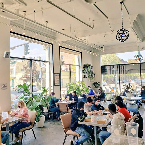 People gather around tables in a bright café with large windows and leafy plants.