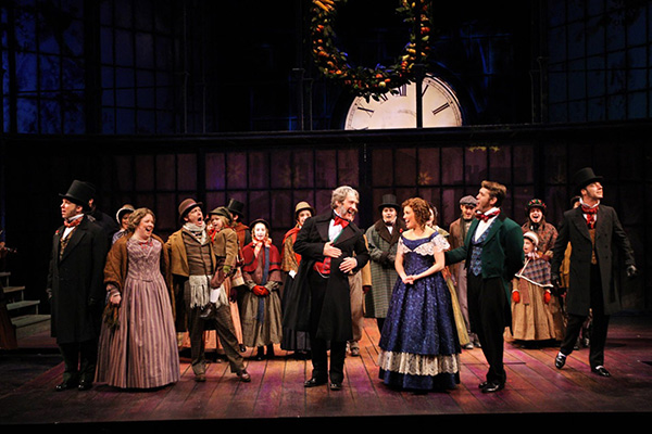 Actors in period costumes perform on a stage beneath a large clock and holiday wreath.