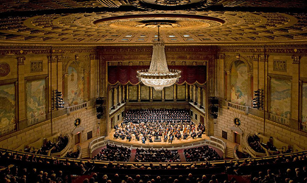 Orchestra and choir performing in an ornate concert hall beneath a chandelier.