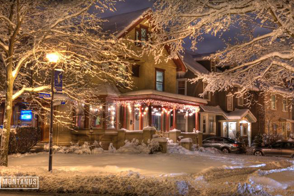 A snow-covered house and trees glow with holiday lights beside a residential street at night.