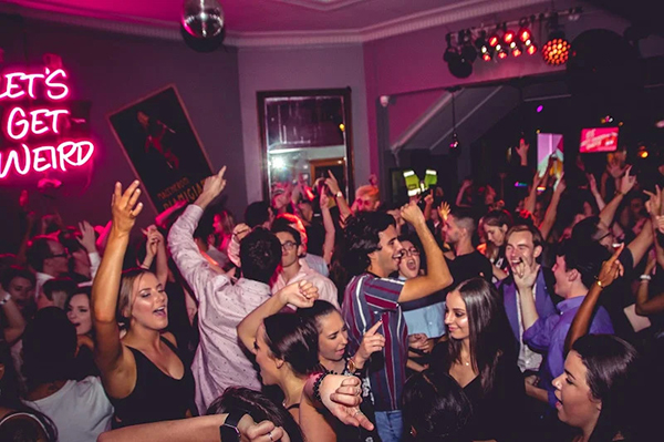 Partygoers dance with raised hands and drinks beneath a disco ball and pink lighting.