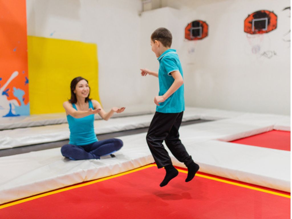 A boy jumps on a trampoline while a smiling woman sits on the padded edge with her arms outstretched.