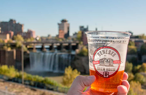 A hand holds a Genesee Brew House beer cup overlooking a waterfall.