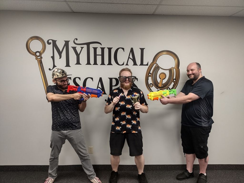 Three people pose with toy blasters, novelty glasses, and a small trophy before a mural.