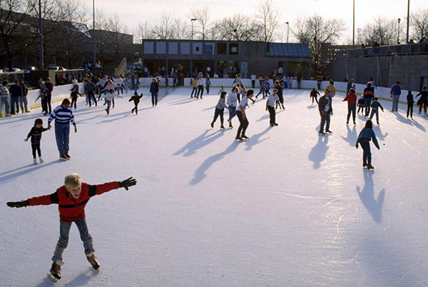 Children and adults ice skating on a sunny outdoor rink.