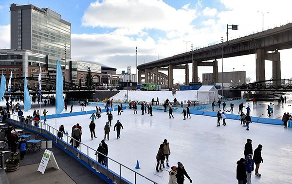 People skating on a large outdoor ice rink beside an elevated roadway
