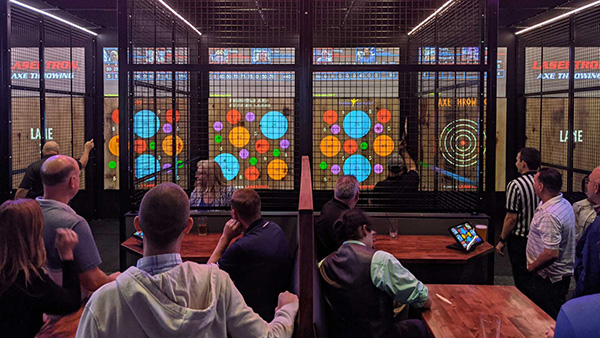 People gathered beside fenced axe-throwing lanes with projected targets.