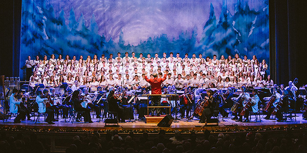 Conductor leading an orchestra and choir before a snowy forest backdrop.