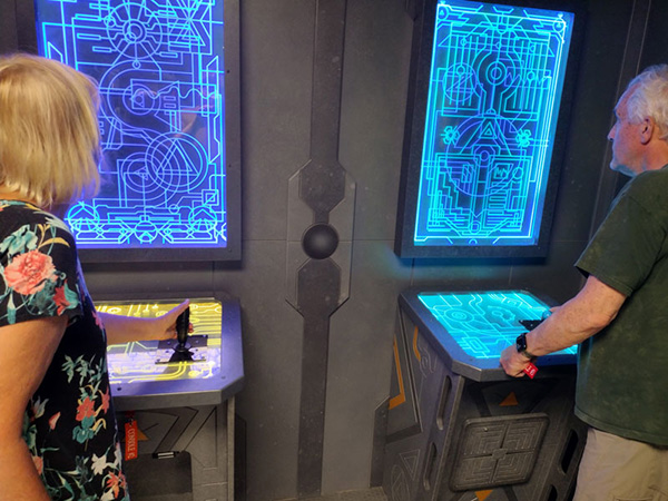 Two adults using glowing control panels beneath blue diagrams in a futuristic room