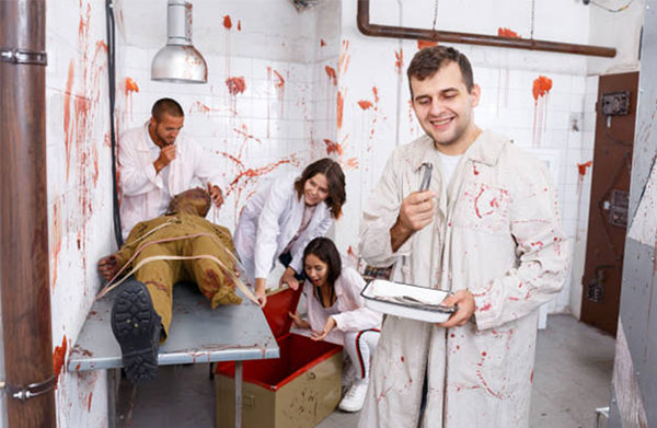 People in red-stained lab coats surround a strapped-down figure in a staged horror laboratory.