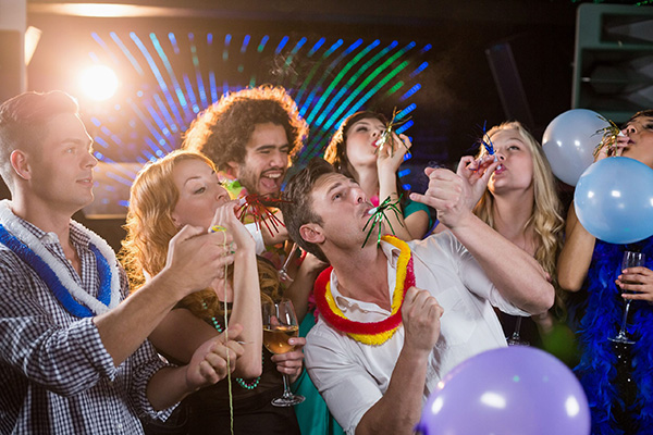 Partygoers blow colorful party horns and hold drinks among balloons and festive leis.