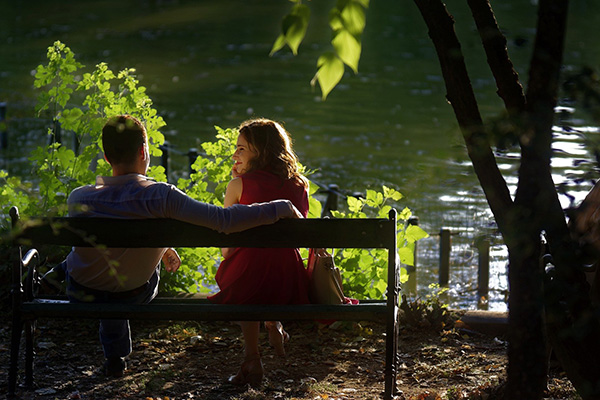 Two people sit together on a park bench beside sunlit water.