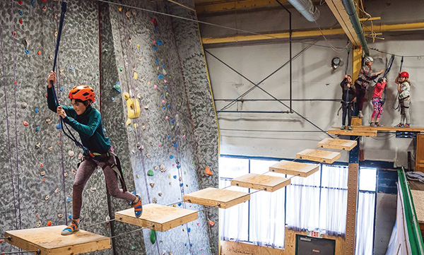 People wearing helmets and safety harnesses cross suspended platforms on an indoor ropes course.