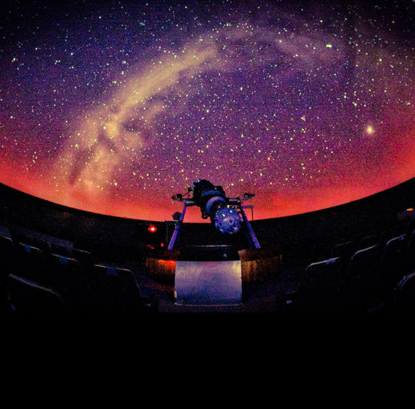A planetarium projector stands beneath a domed display of stars and a glowing purple and orange sky.