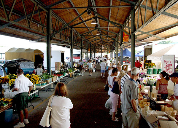 Shoppers browsing produce, flowers and food stalls in a covered market.