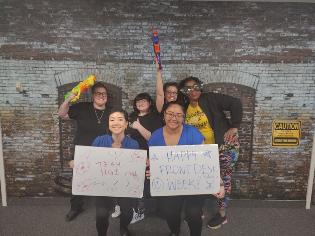 Six people pose with toy blasters and signs reading “TEAM HGI” and “HAPPY FRONT DESK WEEK!”