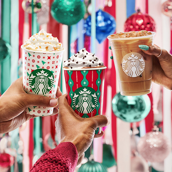 Three hands holding Starbucks drinks in festive cups, including two topped with whipped cream.