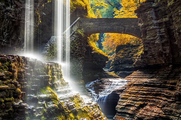 Waterfalls cascading through a rocky gorge beneath an arched stone bridge and autumn trees.