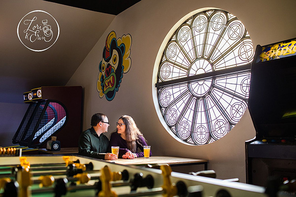 Two people sit with drinks in an arcade space with a foosball table and a large circular window.