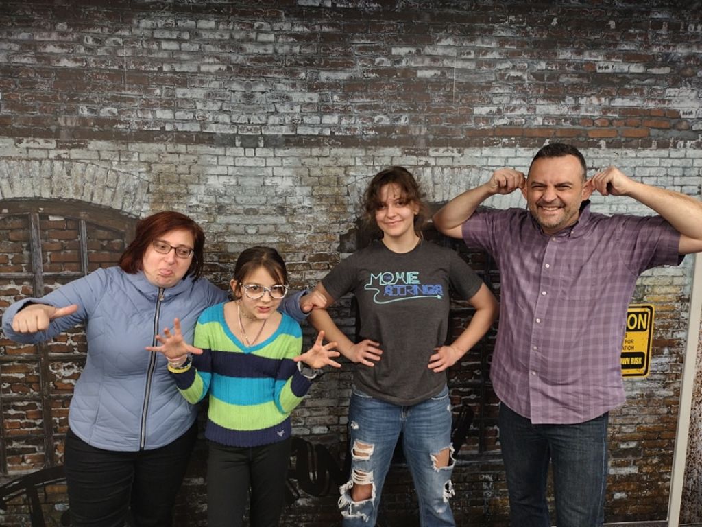 Four people making playful faces and gestures in front of a brick-patterned wall.