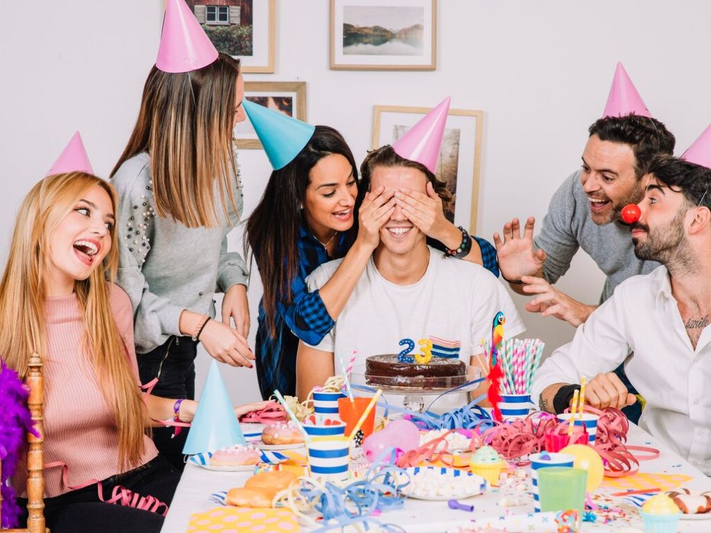 Party guests gather around a cake with “23” candles as one person covers the birthday guest's eyes.