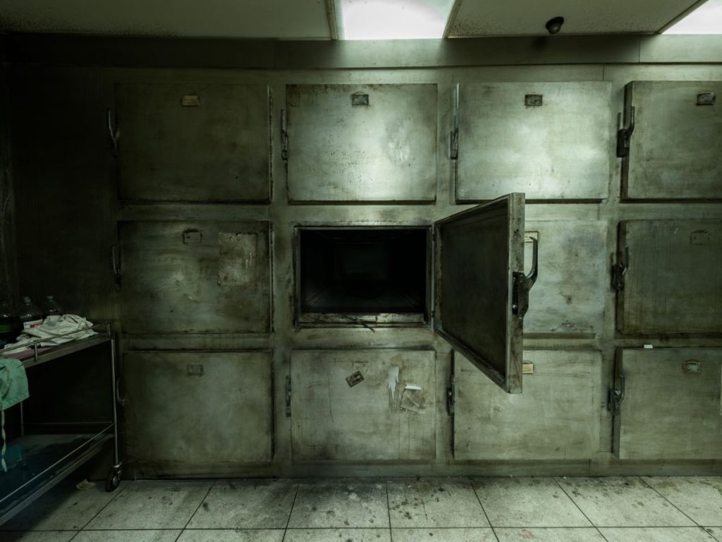 Dim room with metal morgue-style compartments, one open, and a wheeled cart