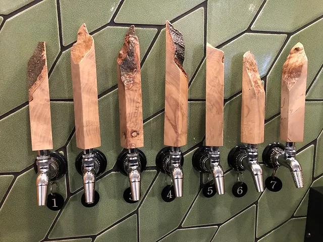 Seven beer taps with tall, irregular wooden handles mounted against a green tiled wall.