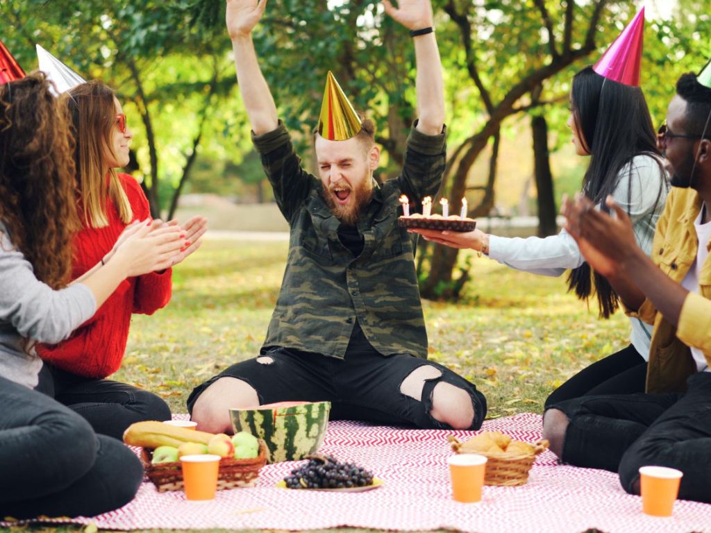 A man in a gold party hat raises his arms as others clap beside a birthday cake at an outdoor picnic.