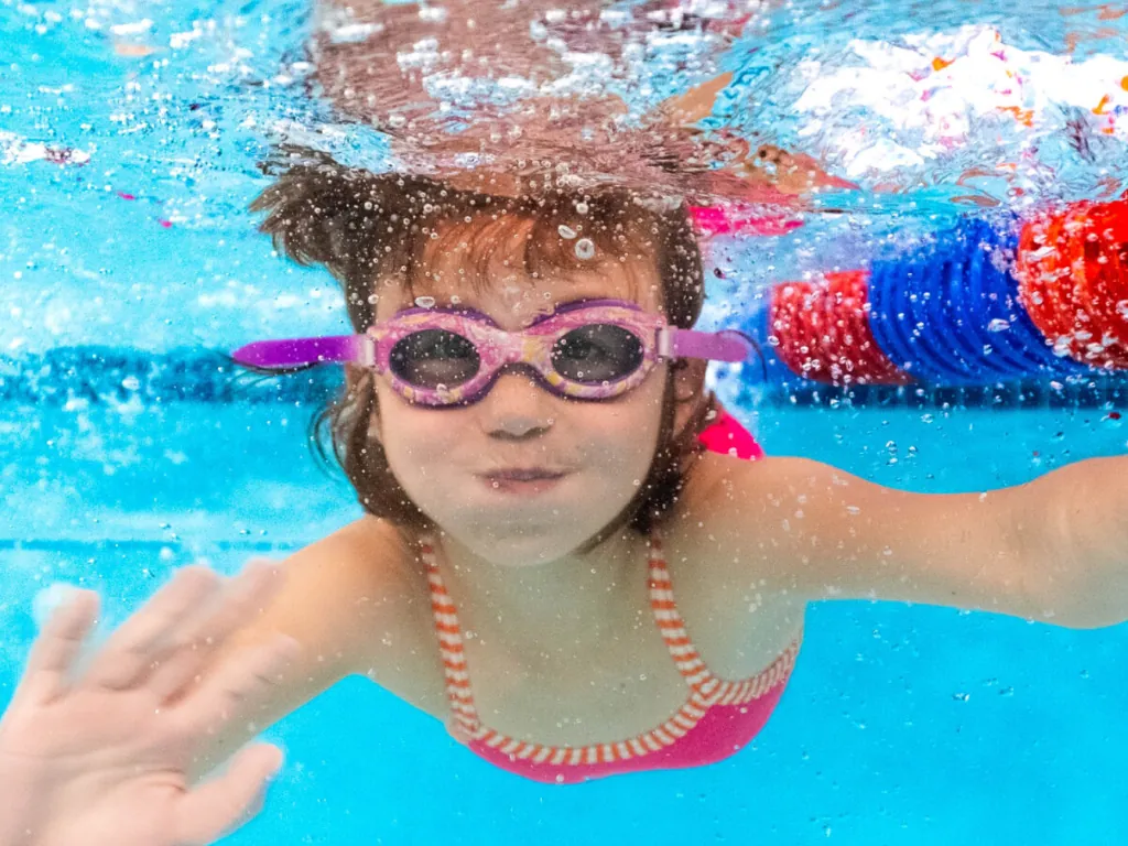 A child wearing purple goggles swims underwater beside pool lane dividers.