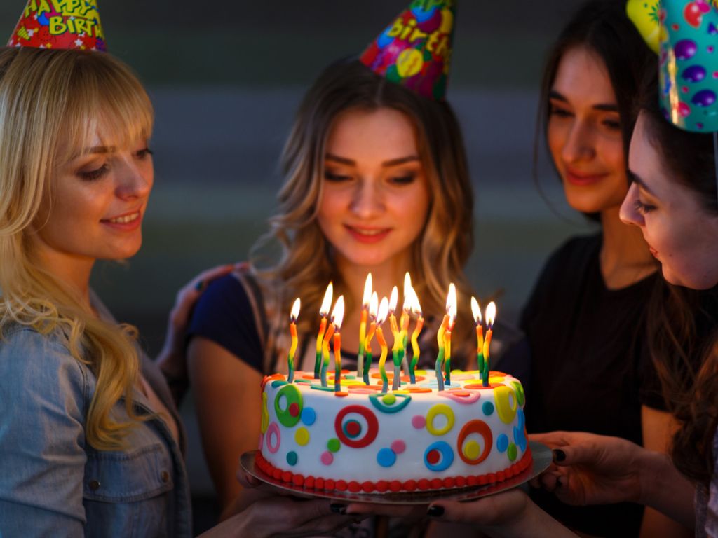 Four women in party hats gathered around a colorful birthday cake with lit candles.