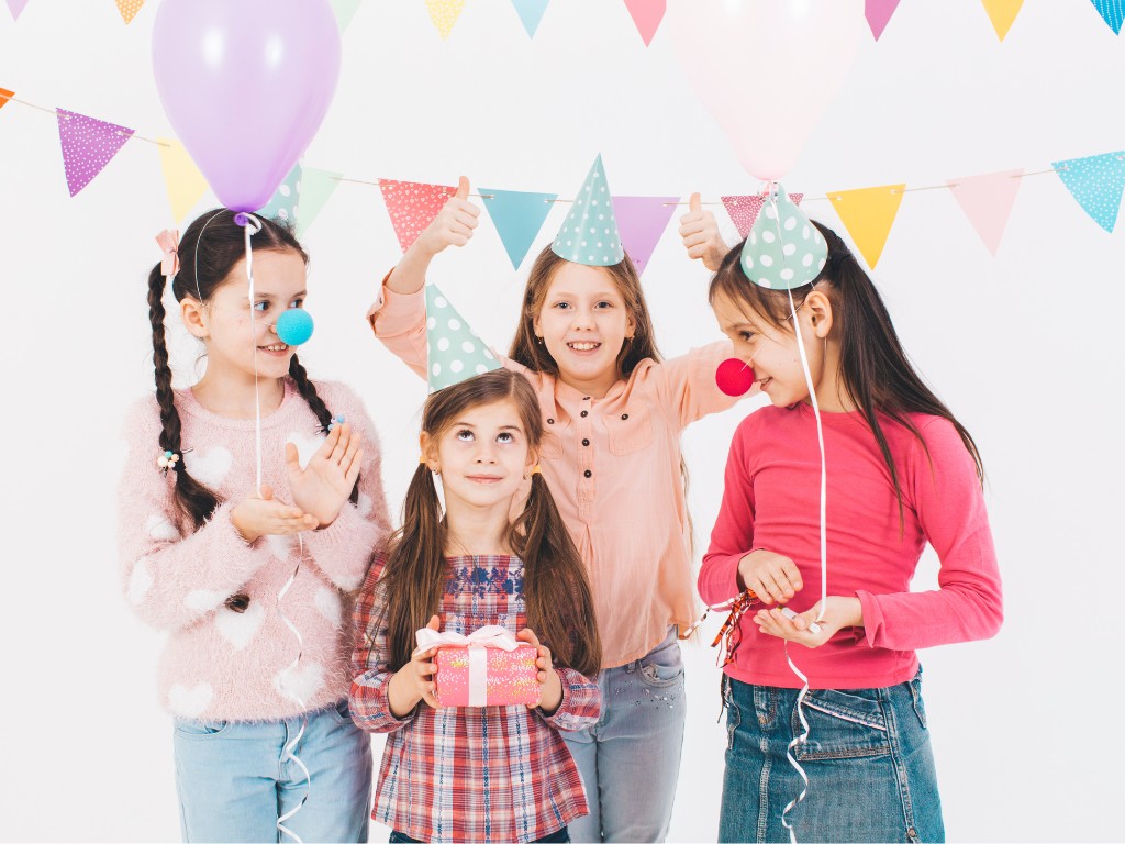 Four children in party hats celebrating with balloons, bunting and a wrapped gift.
