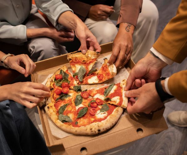 Several people reach into a pizza box and lift slices topped with tomatoes, cheese, and basil.
