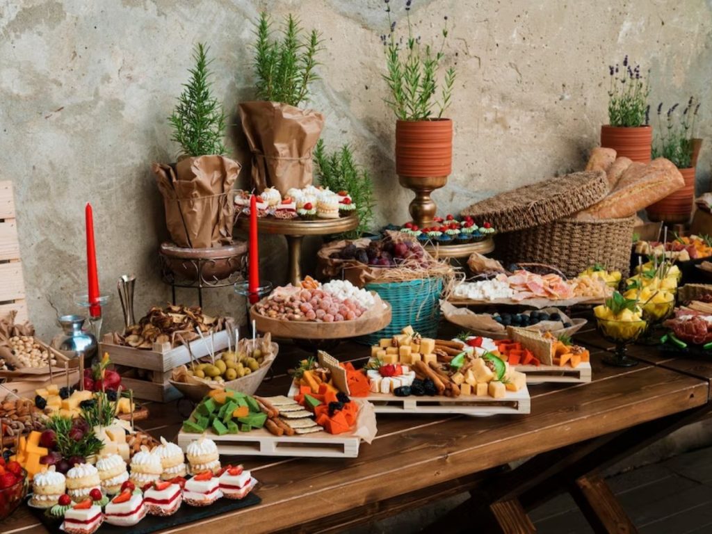 A buffet of cheeses, crackers, olives, cured meats, fruit, baguettes, and small cakes on a rustic table.