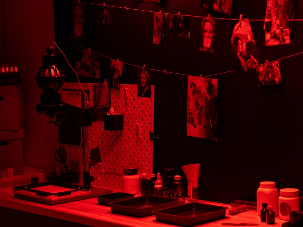 A red-lit photographic darkroom with hanging prints, an enlarger, developing trays, and bottles.