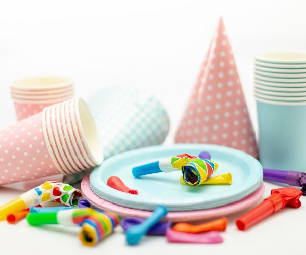 Pastel paper plates, cups and party hats with colorful balloons and party blowers.