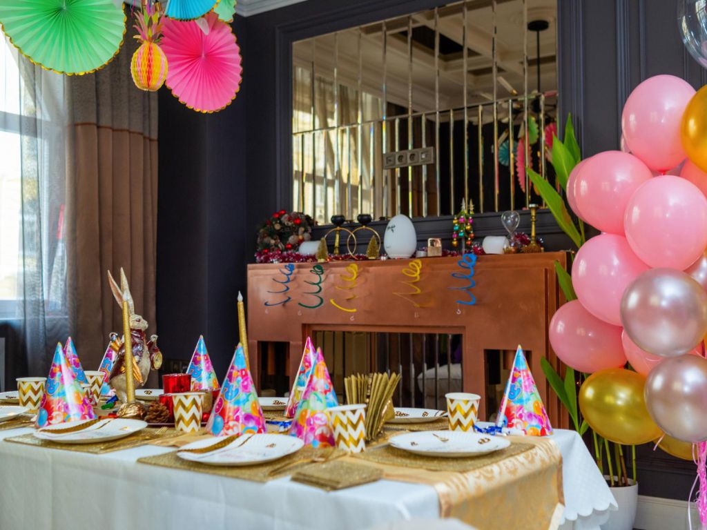 Party table set with colorful hats, gold cutlery and cups, balloons and hanging paper decorations