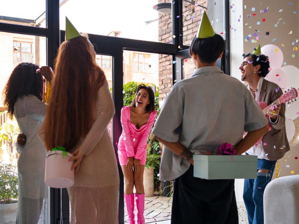 Partygoers greeting a woman in pink with confetti, balloons, gifts and a guitar