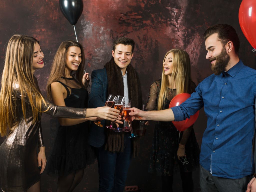 Five people toast with glasses of amber-colored drinks among red and black balloons.