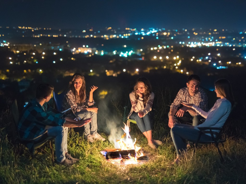 Five people sitting around a campfire at night with city lights in the distance.