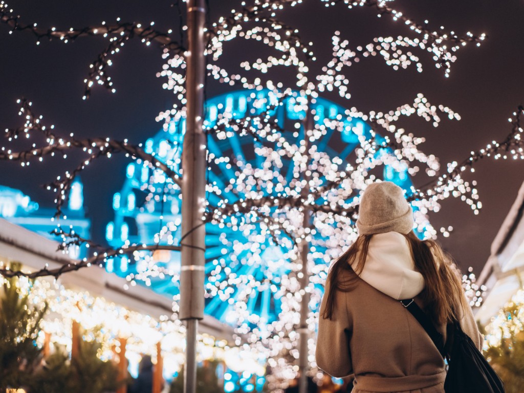 Person beneath illuminated branches at a nighttime market with a glowing Ferris wheel