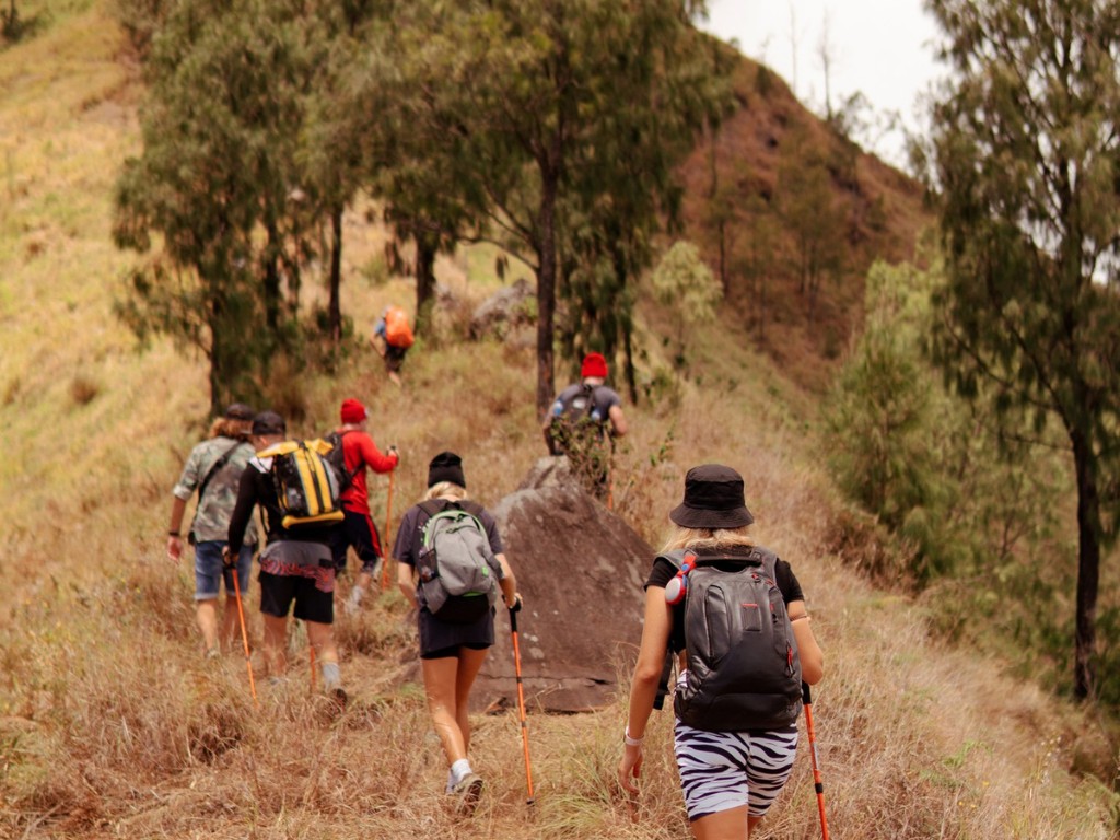 Hikers with backpacks and trekking poles follow an uphill trail through grass and rocks.