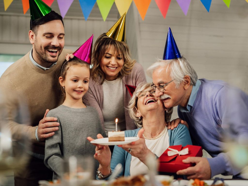 Partygoers in colorful hats celebrating an older woman holding a cake with a lit candle