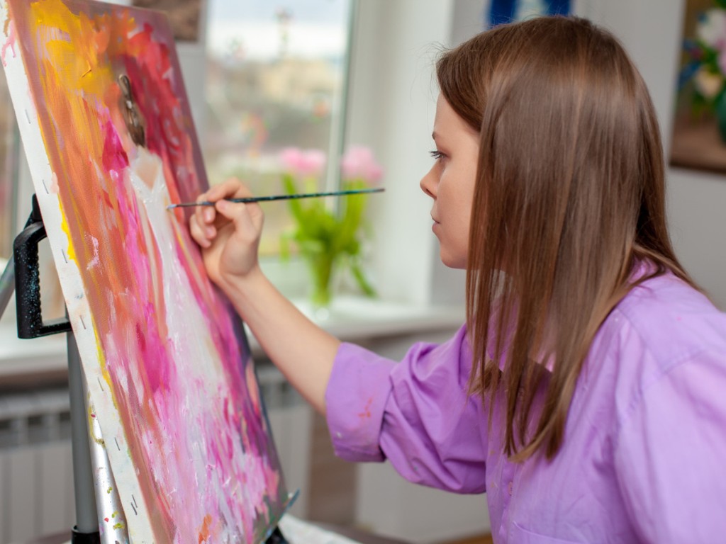 A person uses a fine brush to paint a colorful canvas on an easel.