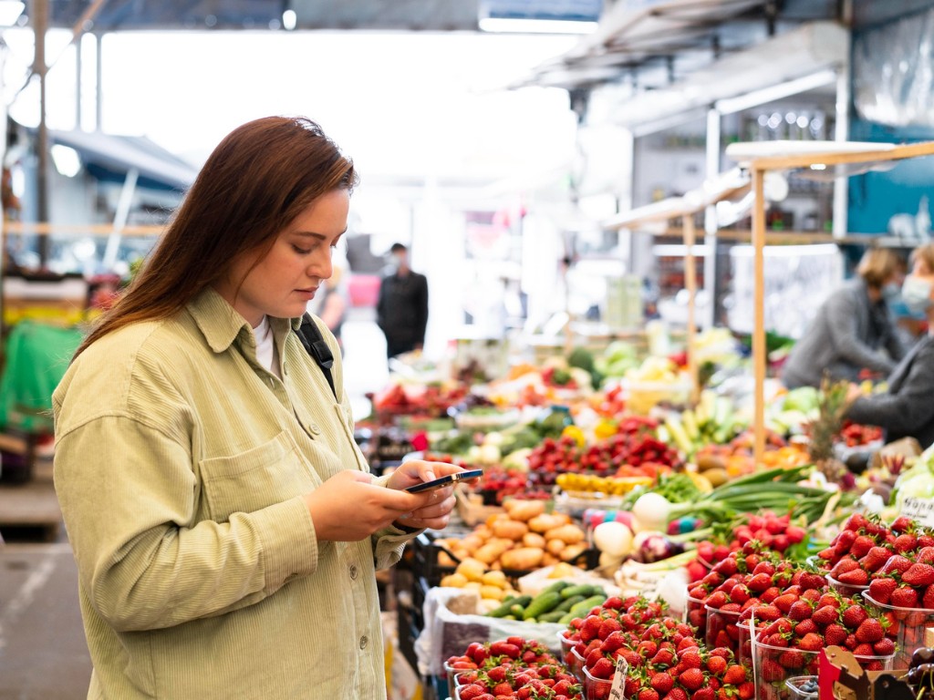 A woman looks at her phone beside a produce stall filled with strawberries and vegetables.