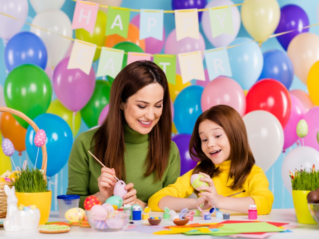 Adult and child decorating Easter eggs with paints beneath a Happy Easter banner