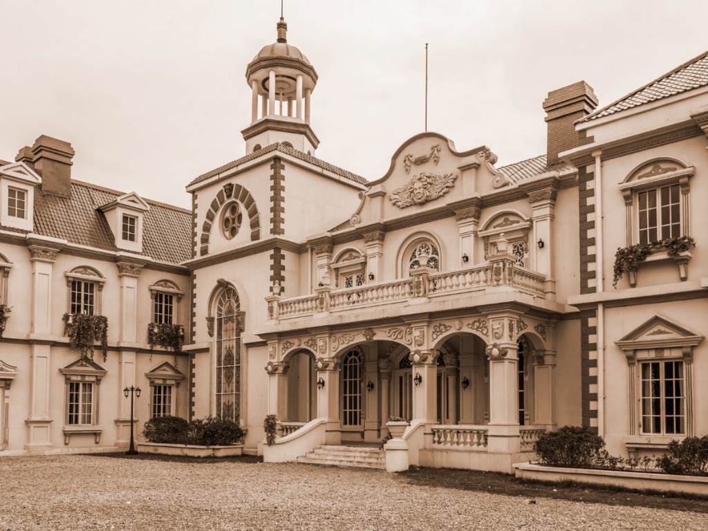 An ornate building with a cupola, arched entrance, and balustraded balcony faces a broad gravel courtyard.