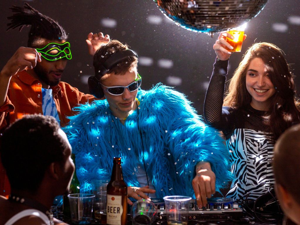 DJ in a bright blue furry coat mixing music as partygoers dance beneath a disco ball