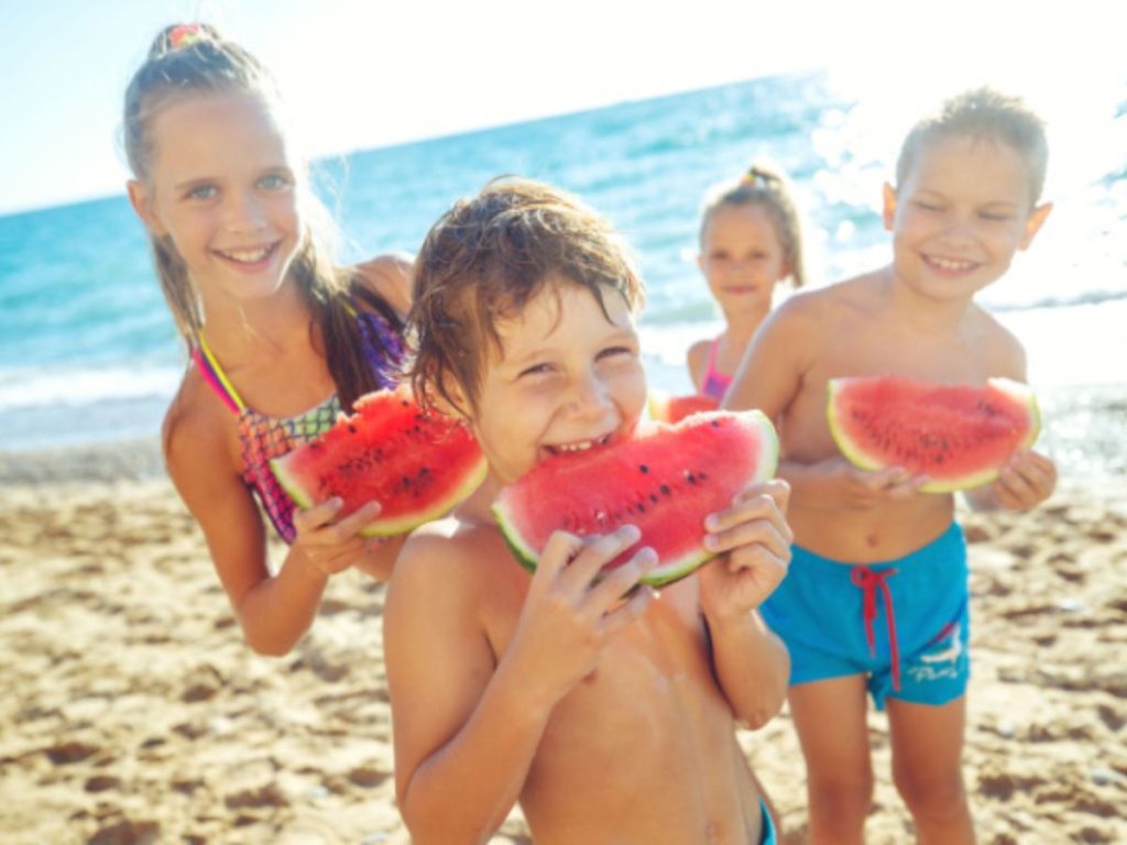 Children in swimwear hold and eat watermelon slices on a sandy beach.