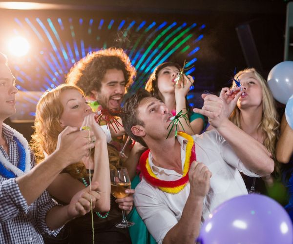 Partygoers blowing party horns among balloons and colorful lights.