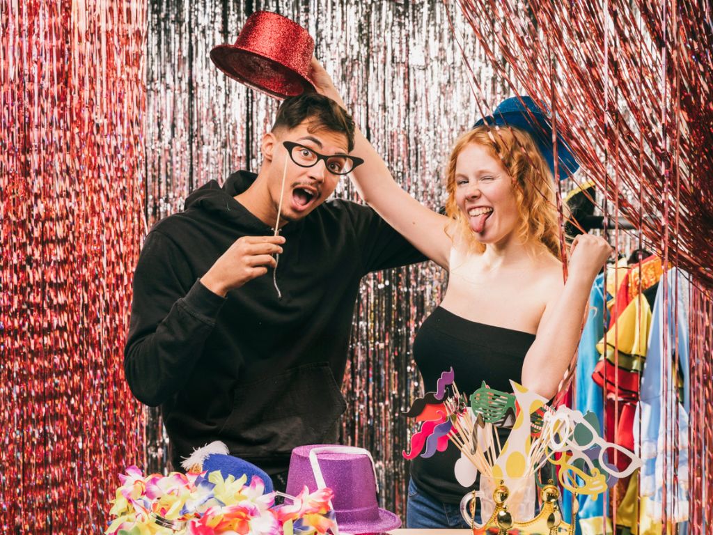 Two people posing with novelty glasses and a red top hat in front of tinsel curtains and a table of photo props